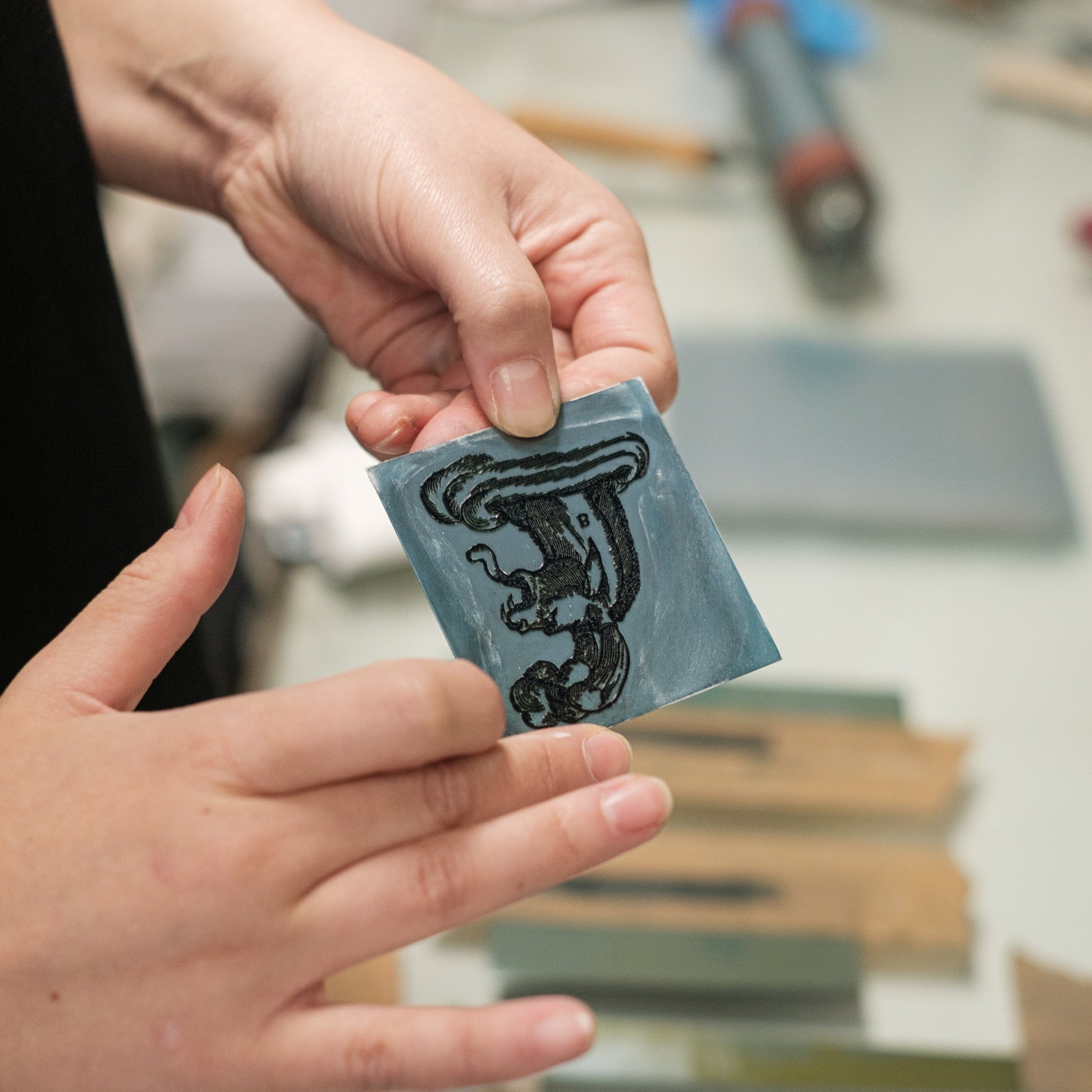 Printmaker holding a small blue printing plate with the image of a monster design, on a blurred background of tools and materials